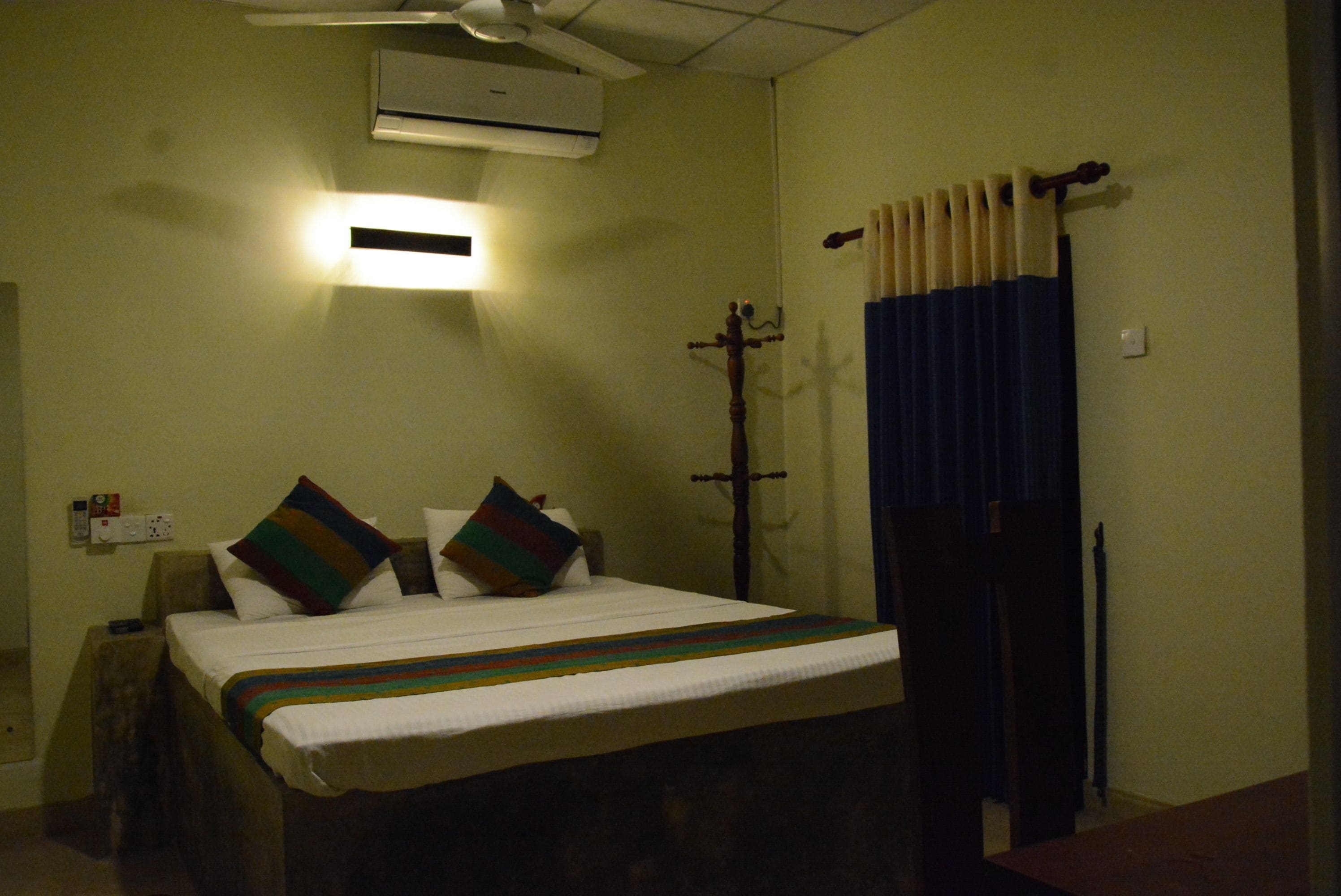 A hotel room with a bed, a ceiling fan, an air conditioner, a nightstand, and a window with curtains.