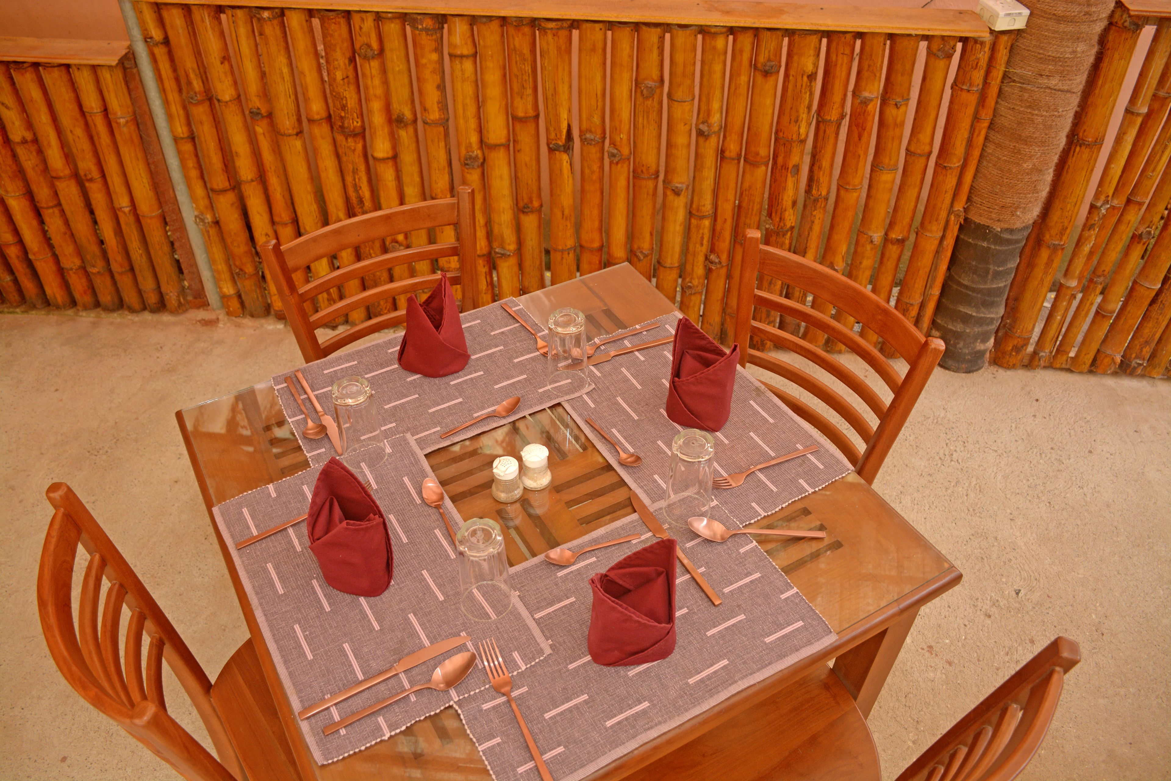 Una mesa cuadrada con cubierta de vidrio, rodeada por cuatro sillas de madera. La mesa está dispuesta con cubiertos, vasos y servilletas.