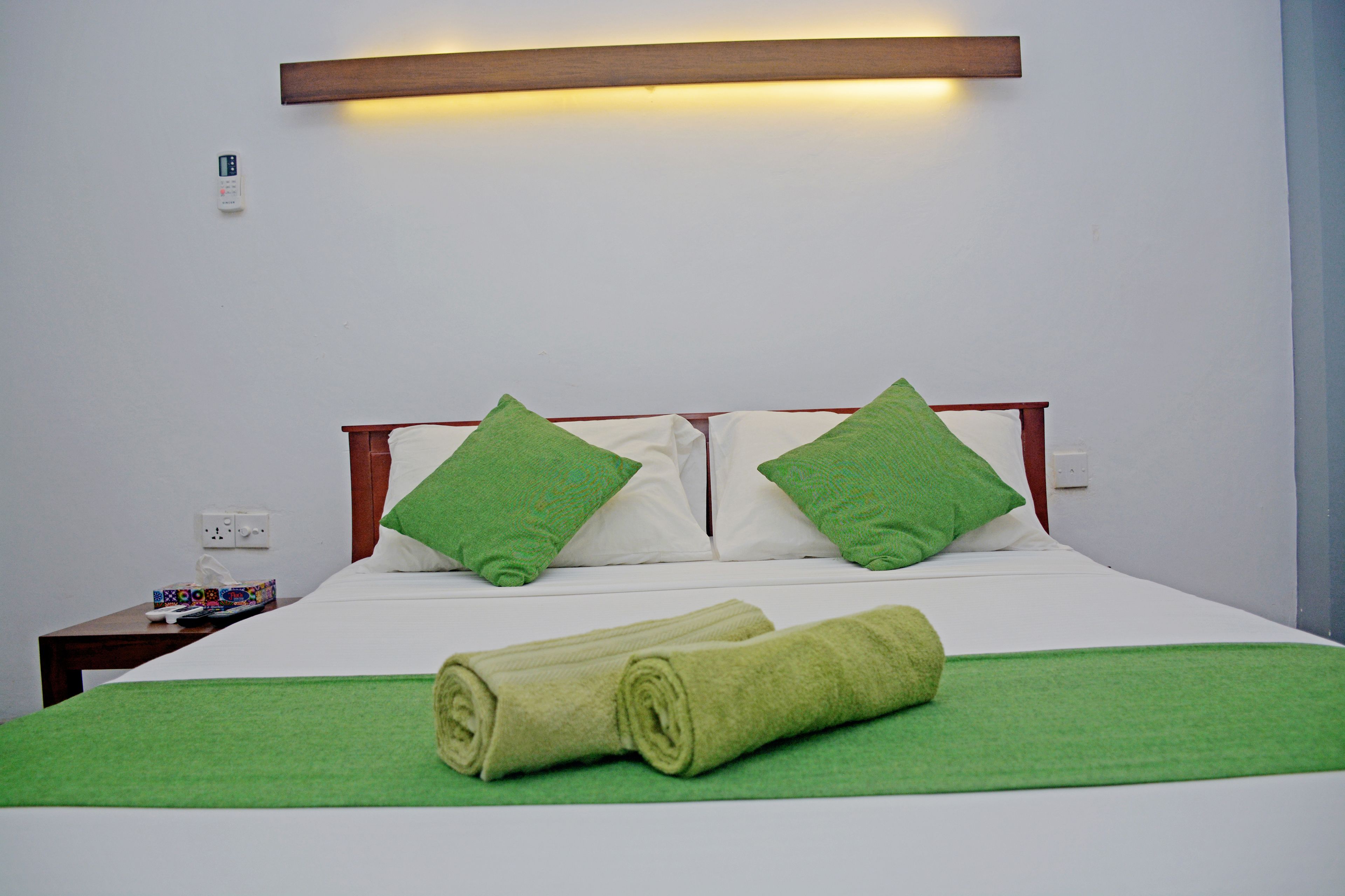 Una cama bien hecha con toallas y almohadas verdes, una mesita de noche con control remoto y una lámpara fijada a la pared.