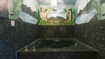 Una bañera grande y separada en un cuarto con un mural de paisajes tropicales y un espejo grande encima.