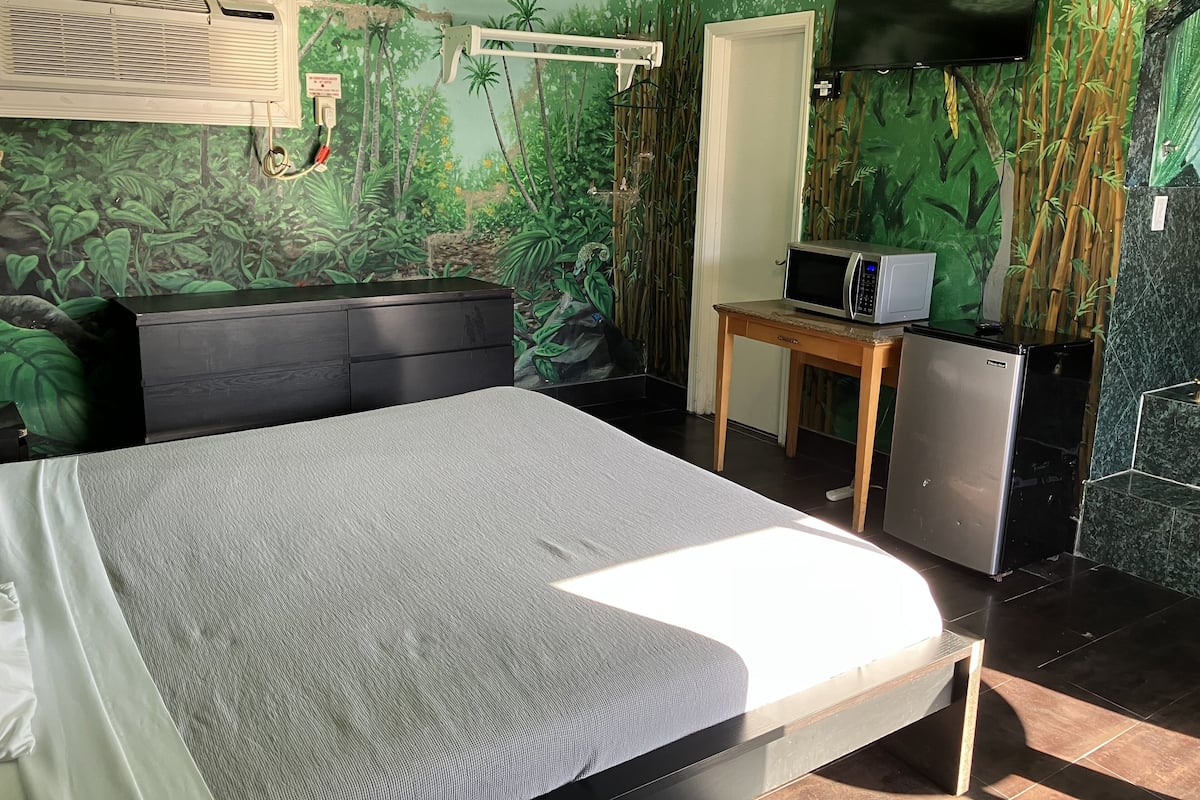 A bedroom with a bed, a desk with a microwave, a refrigerator, and a TV mounted on the wall.