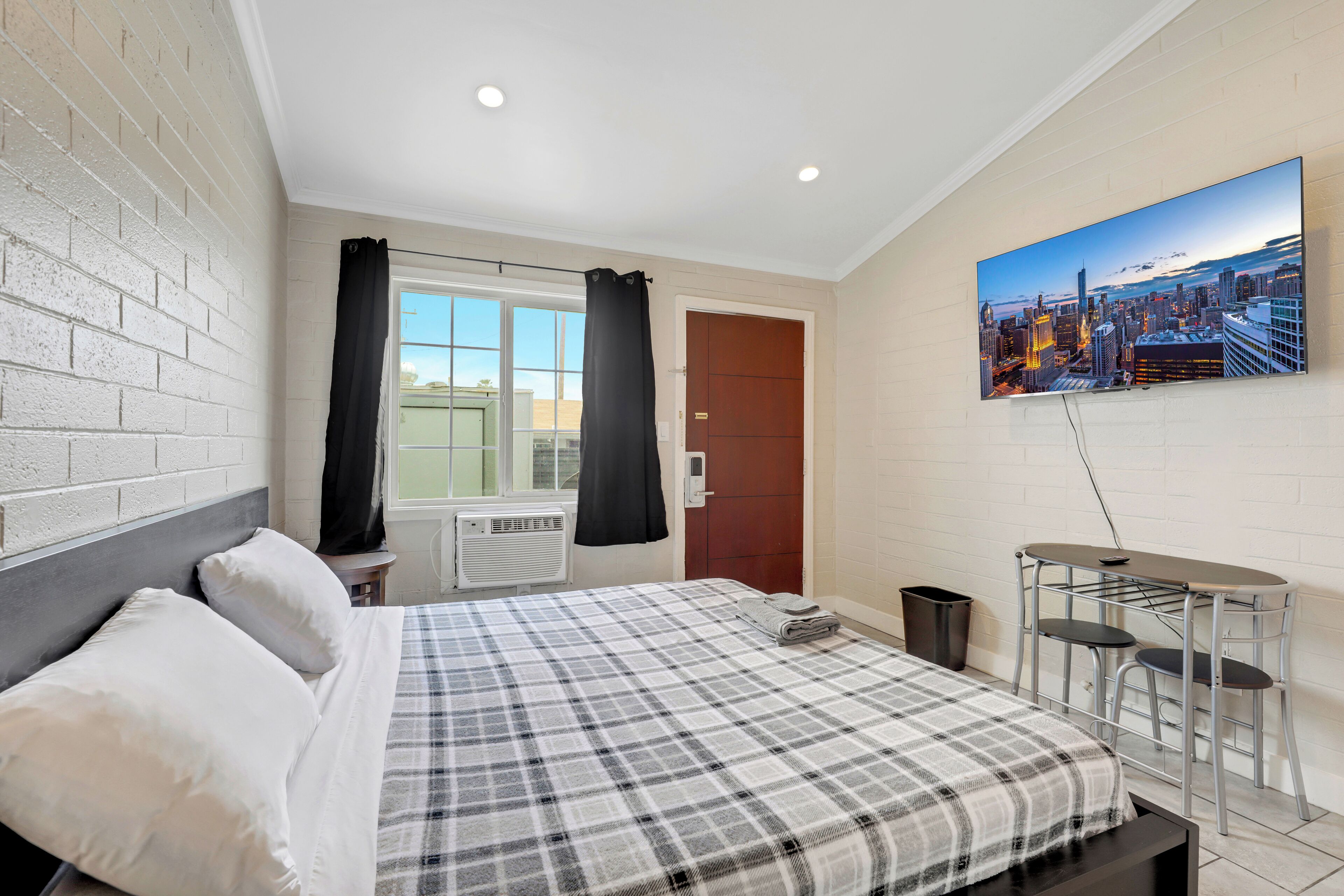 A bedroom with a bed, a window with curtains, a door, a small table with chairs, and a cityscape painting on the wall.