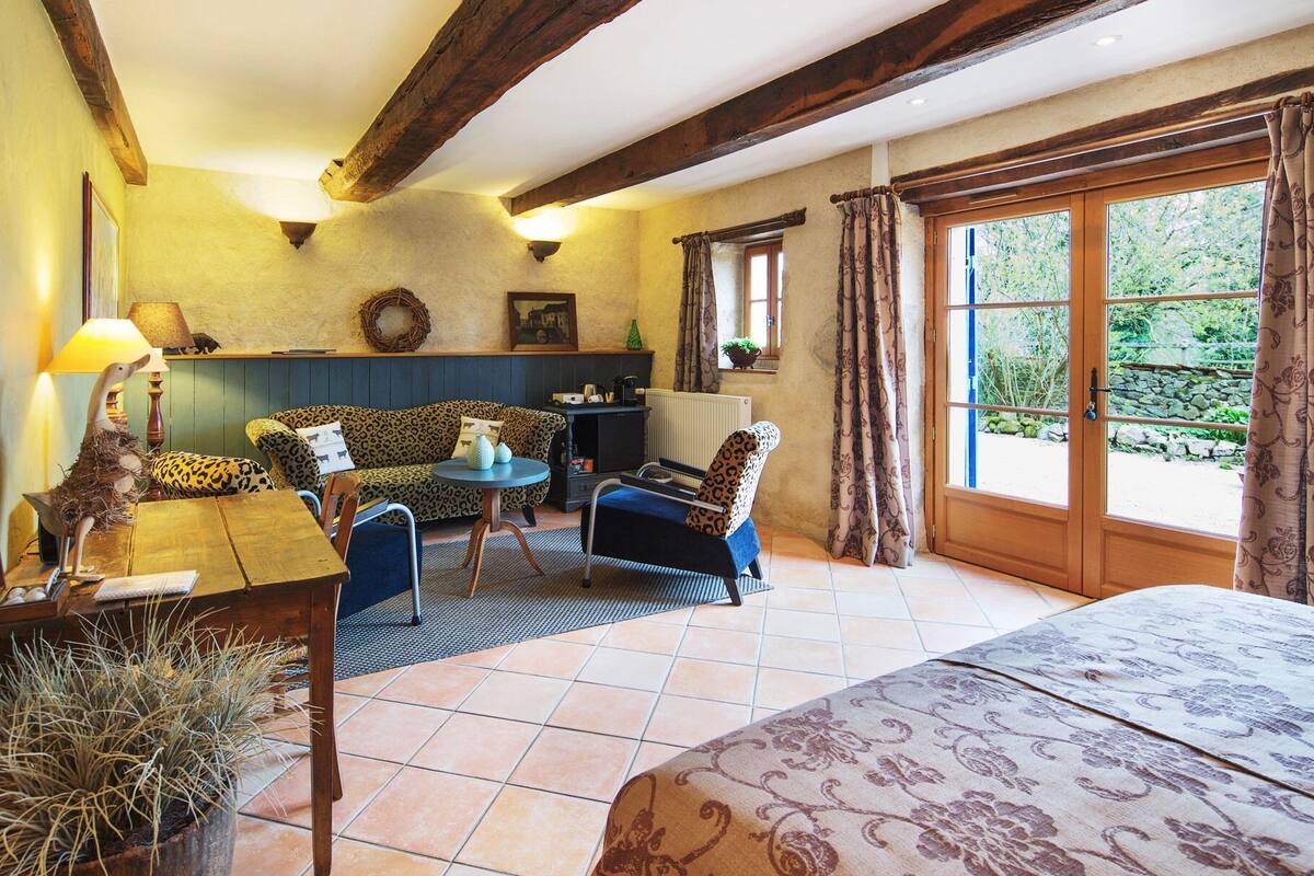 Ein gemütliches Zimmer mit einer Couch, Sesseln, einem Tisch und einem Bett. Ein Fenster mit Vorhängen bietet Blick auf eine grüne Landschaft.