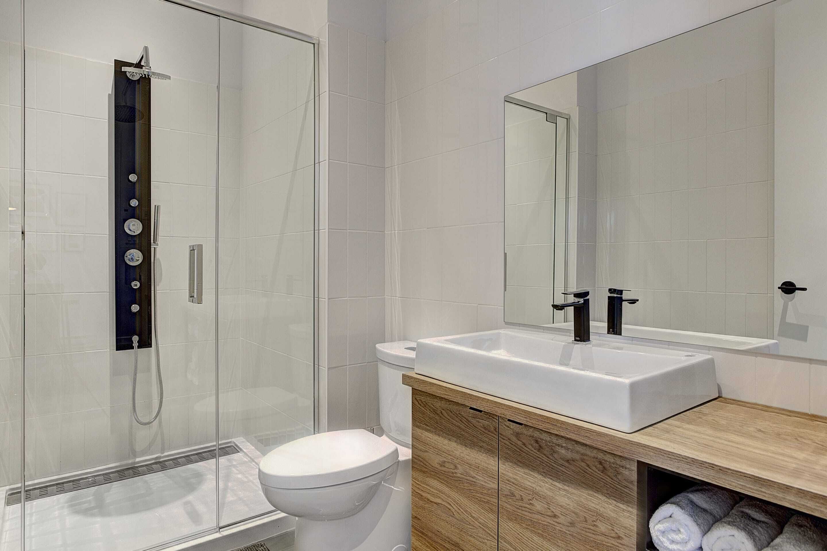 A modern bathroom with a glass-enclosed shower, a white toilet, a wooden vanity with a sink, and a large mirror.