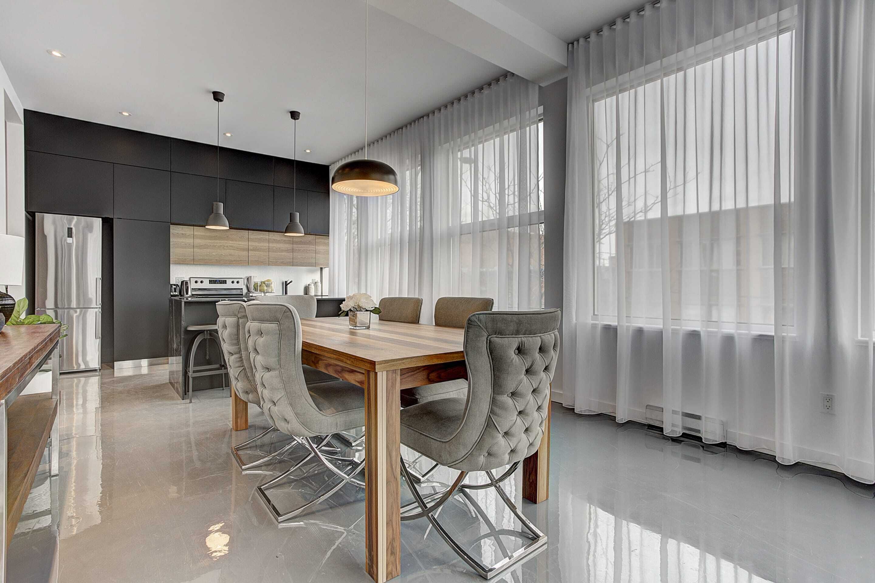 A modern kitchen with dark cabinetry, a wooden dining table, and a large window with sheer curtains.
