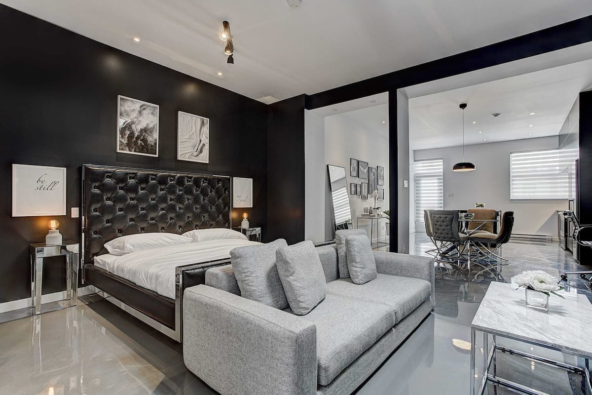 A modern bedroom with a large bed, a grey sofa, and a dining area with a table and chairs.