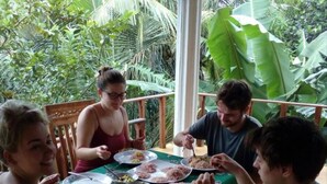 Een groep mensen dineert aan een buitentafel met een groene tafelkleed, omgeven door weelderige tropische beplanting.