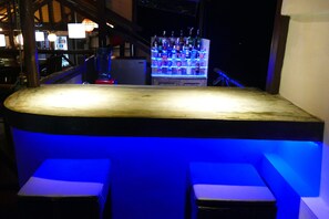 A bar area with a counter and two seating stools, illuminated by blue lighting.