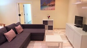 A living room with a dark grey sectional sofa, a white coffee table, a TV on a white cabinet, and a painting of a beach scene on the wall.