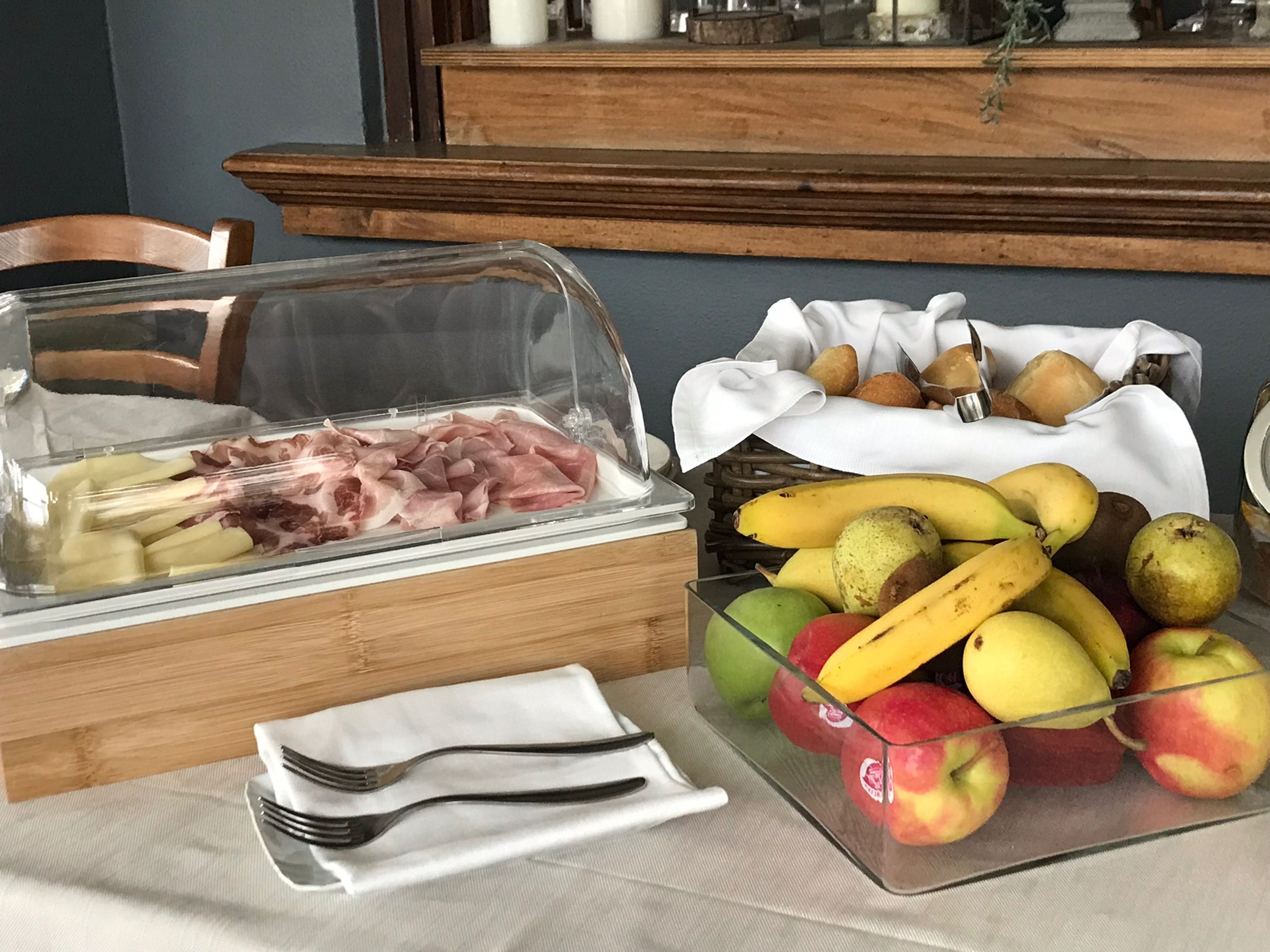 Un buffet con un contenitore di vetro pieno di salumi, una ciotola di frutta assortita tra cui banane e mele, e un cestino di pane.