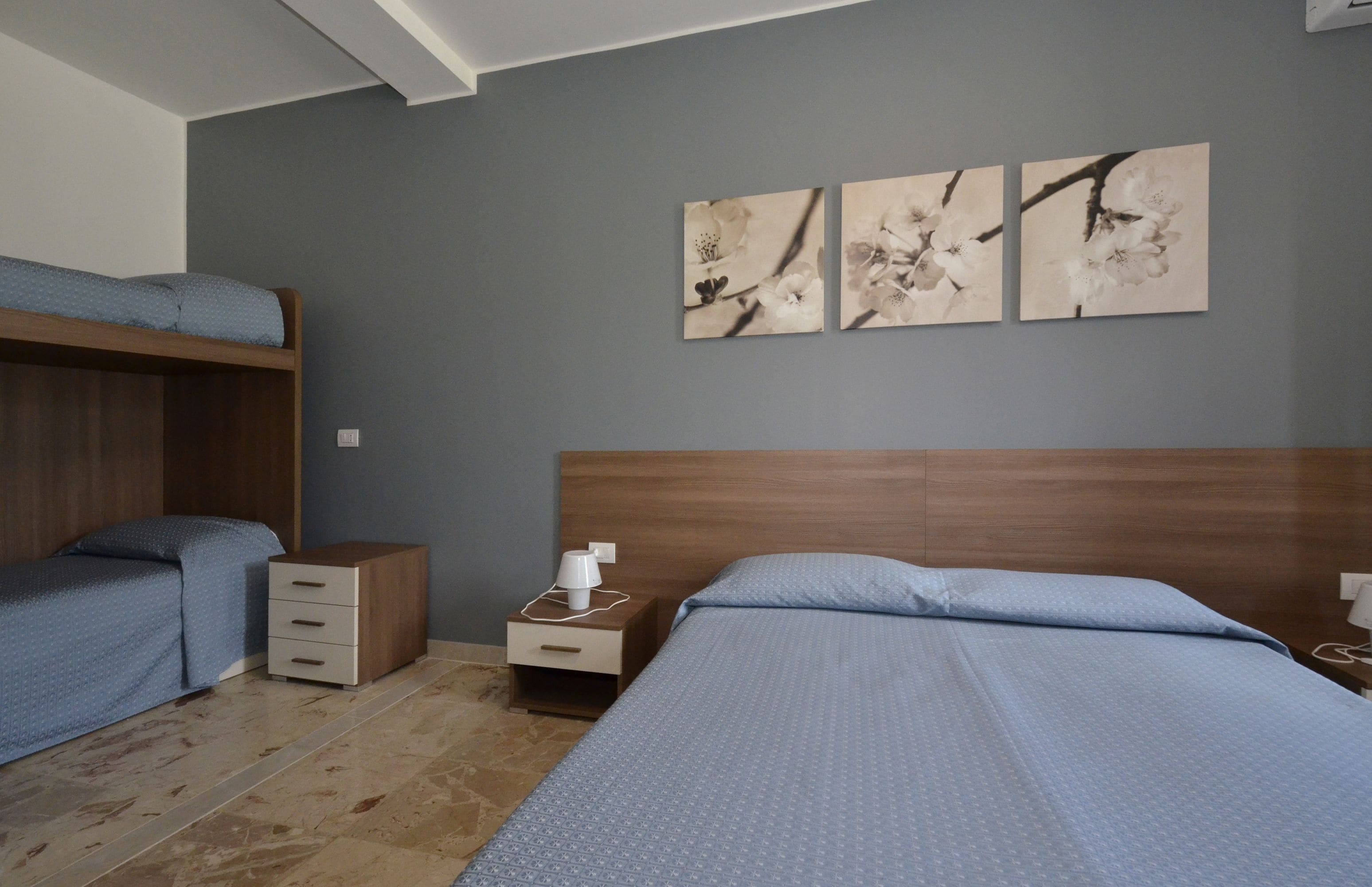 A bedroom with a bunk bed, a single bed, a nightstand with a lamp, and three wall-mounted flower paintings.