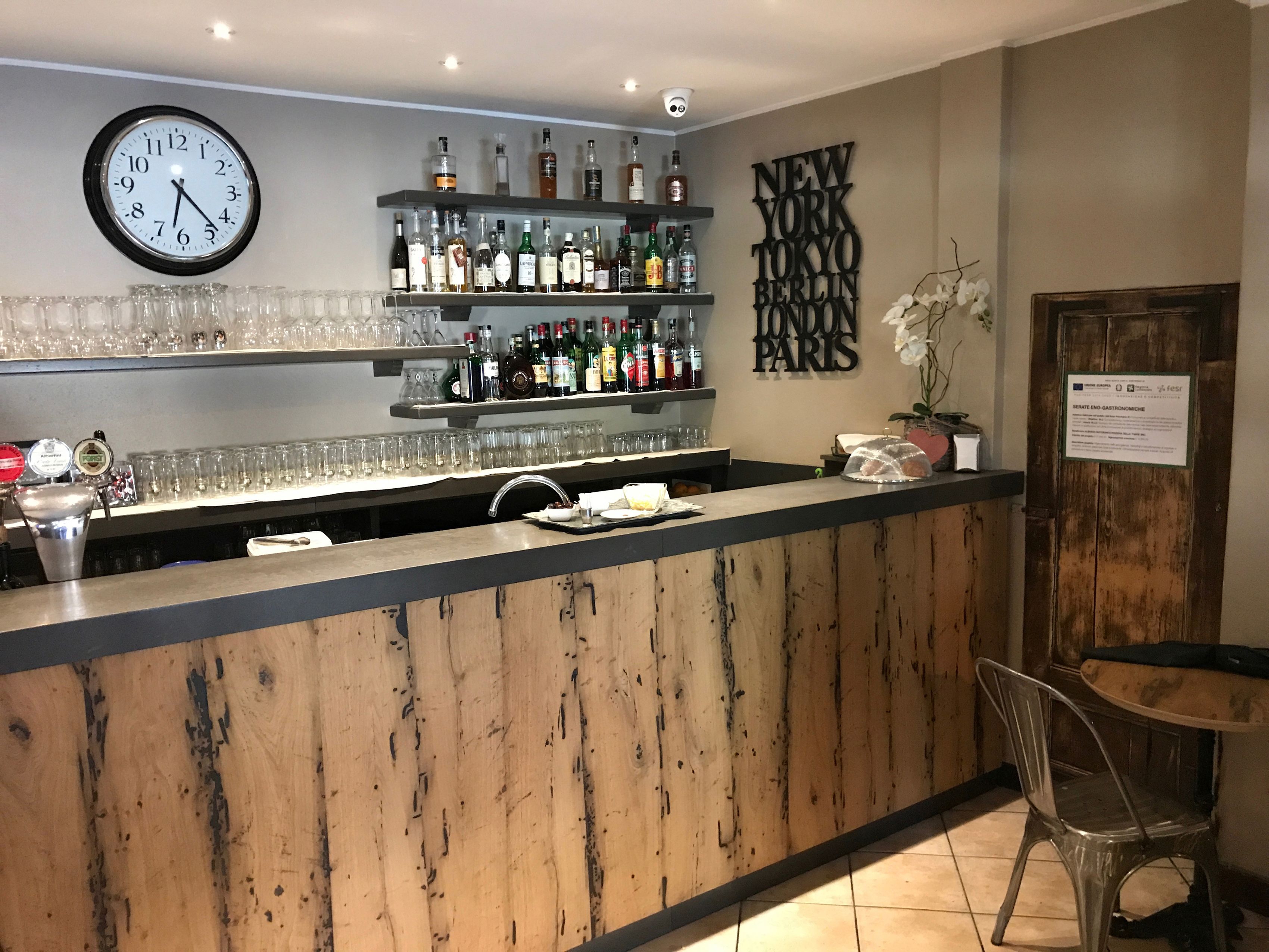 Una zona de bar con mostrador de madera, estantes con botellas, vasos y un reloj.