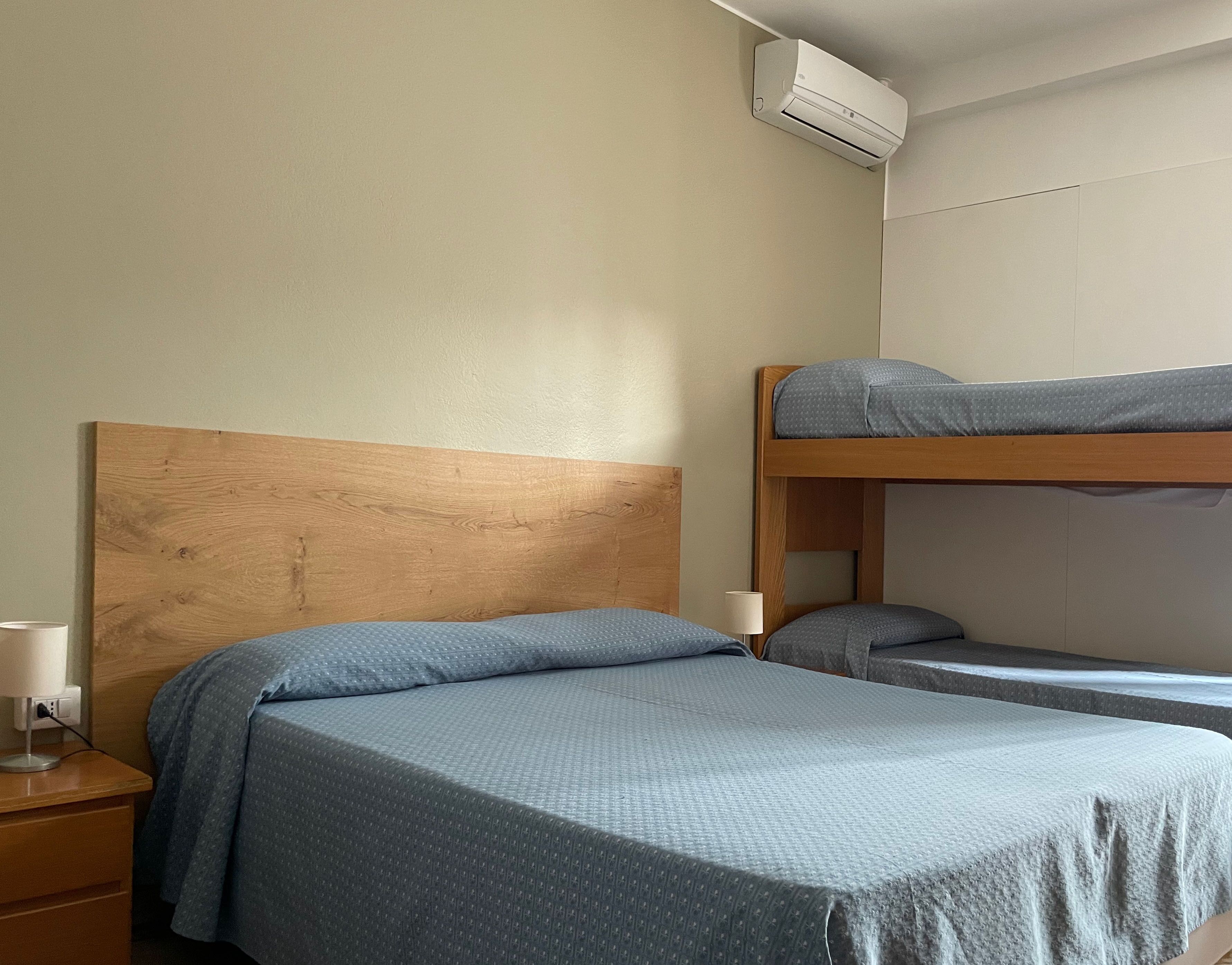 Habitació amb un llit superposat, un llit individual i una tauleta de nit amb una làmpada.