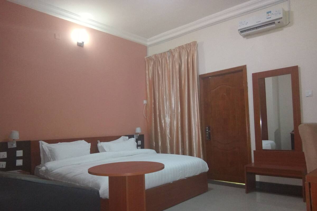 A hotel room with a bed, a round wooden table, a chair, a mirror, and an air conditioning unit.