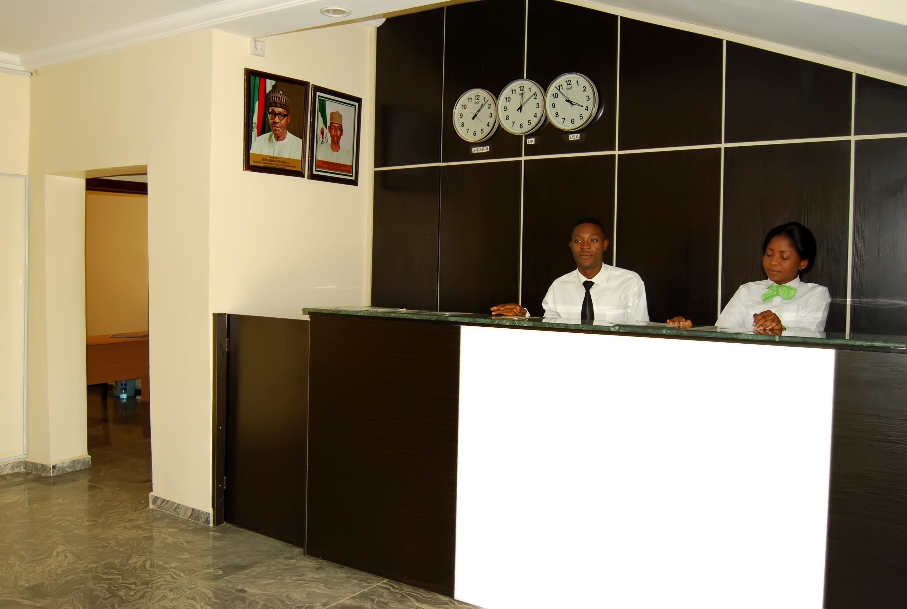 A reception desk with two individuals, a clock, and two framed pictures on the wall.