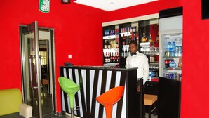 A bar area with a red wall, a refrigerator stocked with beverages, and two colorful bar stools. - Kifina Emerald Hotel (Abuja)