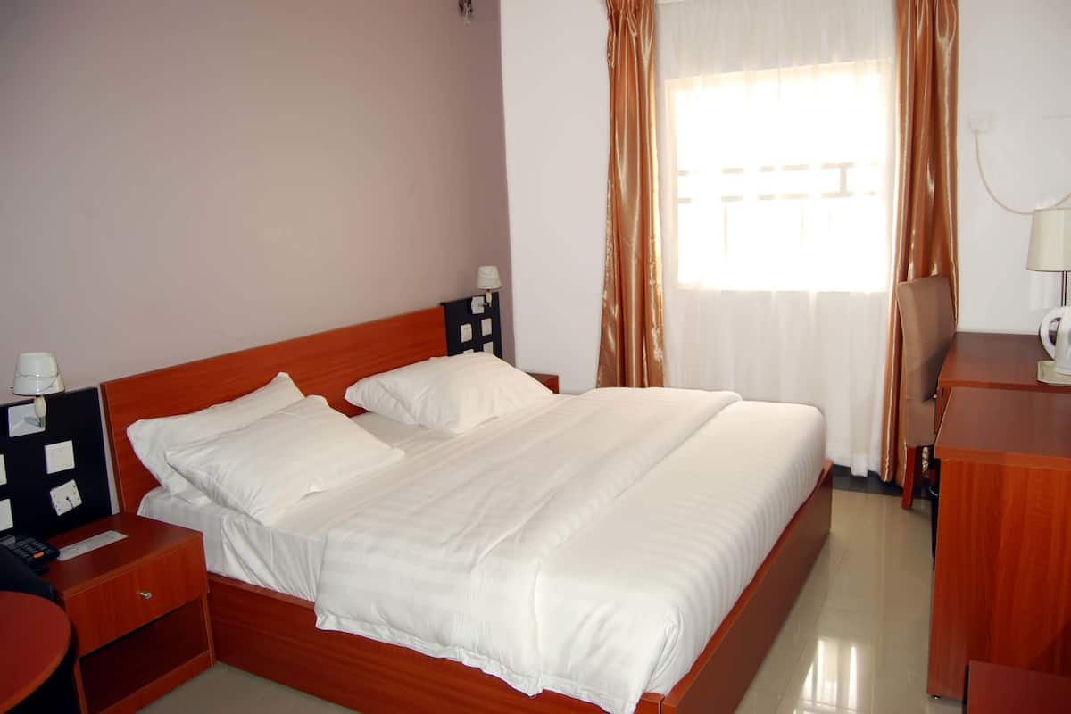 A neatly made bed with white linens, a wooden headboard, a bedside table with a lamp, and a desk with a chair.