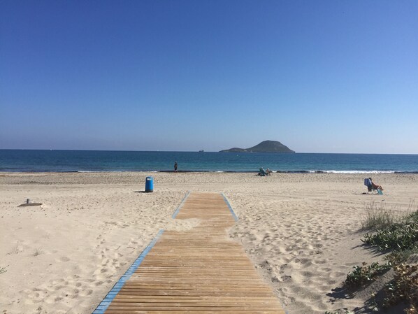 A sandy beach with a wooden boardwalk leading to the water, a small island in the distance, and a trash bin.