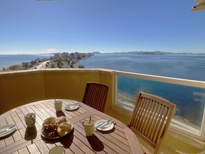 A balcony with a table set for breakfast, overlooking a coastal view with buildings and mountains in the distance. - Front line with stunning sea views! (La Manga del Mar Menor)