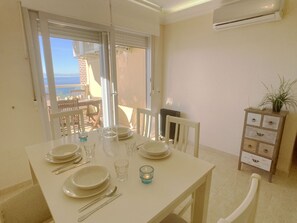 A dining area with a table set for four, a radiator, a wooden cabinet with drawers, and a view of the sea through a window.