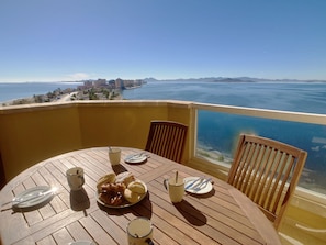A round wooden table with plates, cutlery, and a mug, set against a yellow wall overlooking a coastal cityscape.