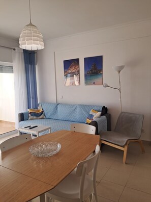 A modern living room with a wooden dining table, a sofa, and a floor lamp. There are two framed pictures on the wall. - Praiamar 3f- Sea Front, Cozy, Private Parking (Quarteira)