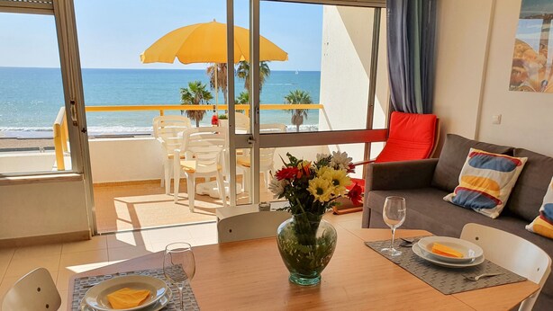 Un coin repas avec une table dressée pour deux personnes, un vase rempli de fleurs et une vue sur l’océan grâce à de grandes fenêtres.