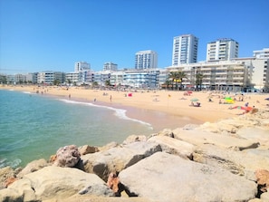 A beach with a rocky foreground, a sandy shore, and a row of multi-story buildings in the background.