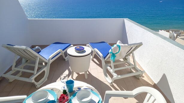 Una terrazza con arredi bianchi, un tavolo apparecchiato con piatti, ciotole e un bicchiere d'acqua, con vista sul mare.