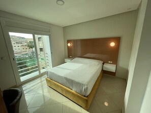 A bedroom with a large bed, a wooden headboard, two bedside lamps, a sliding glass door to a balcony, and a tiled floor. - Puerto Marino (Al Hoceima)