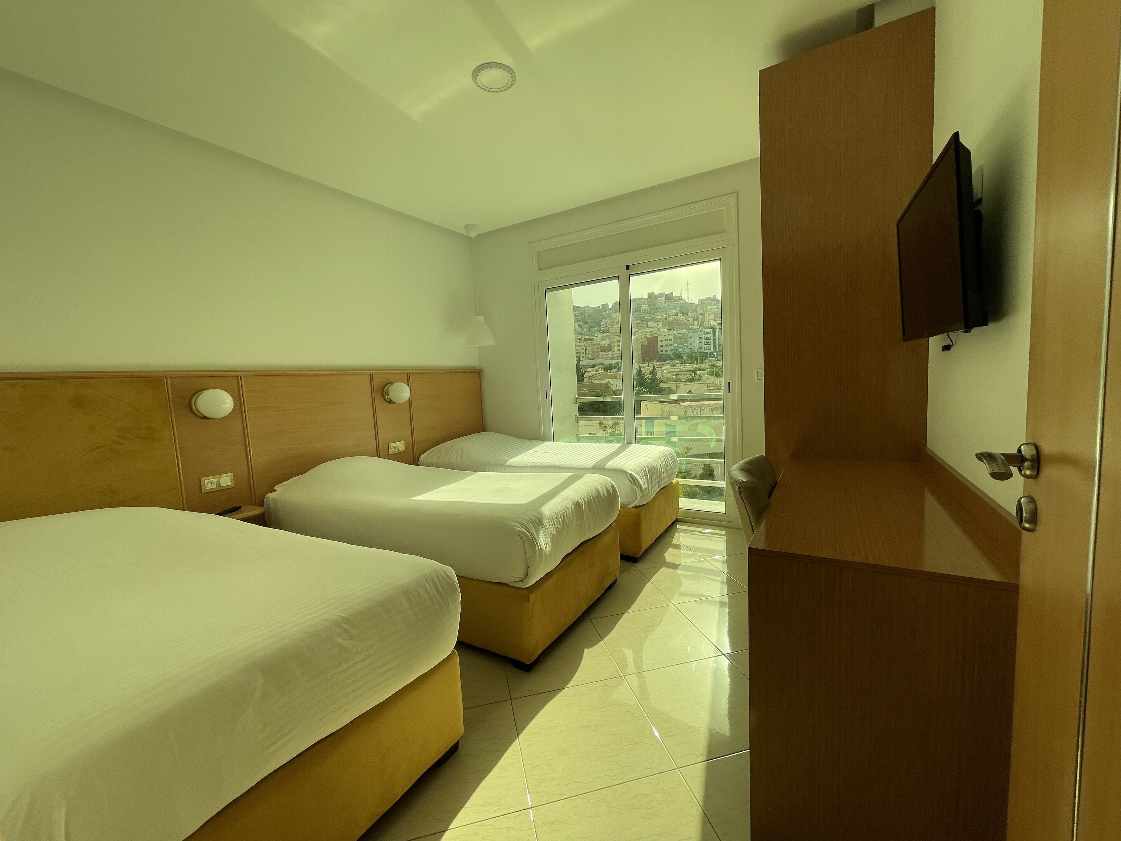 A hotel room with two beds, a television on the wall, and a view of the city through the window.