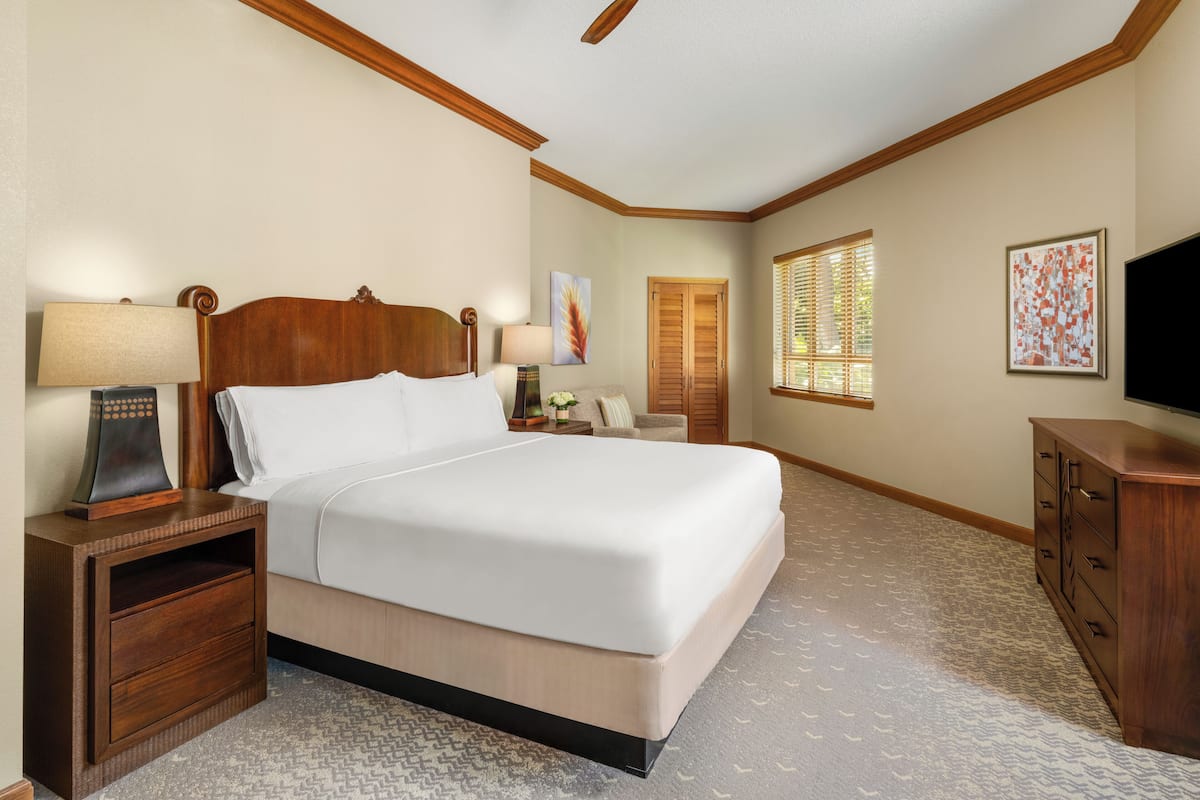 A hotel room with a large bed, wooden headboard, two bedside tables with lamps, a nightstand, a dresser, a ceiling fan, a window with blinds, and a framed picture on the wall.
