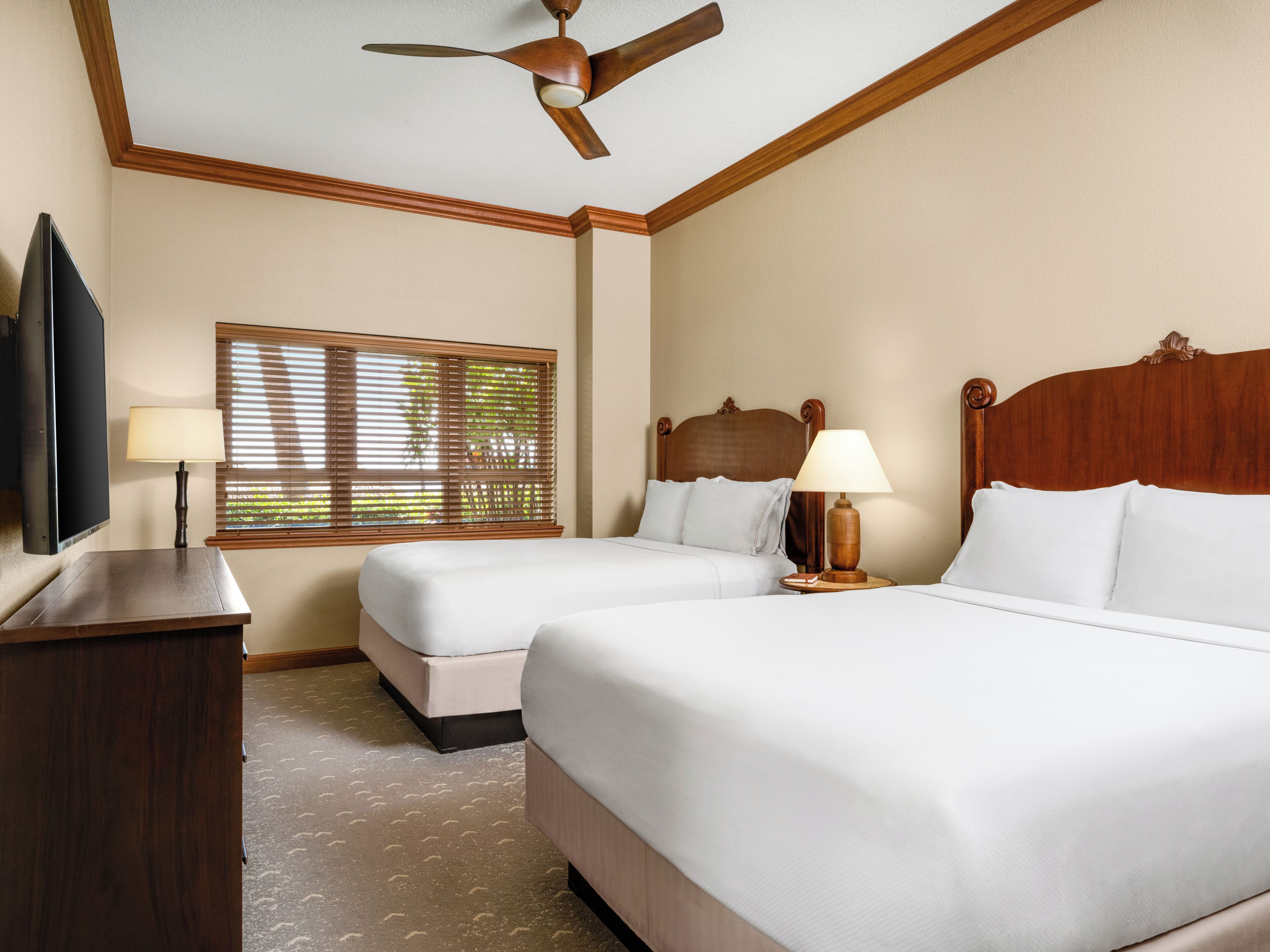A hotel room with two beds, a ceiling fan, a TV, and a window with blinds.