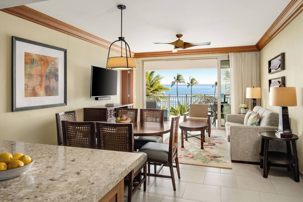 A hotel room with a dining area, a TV, a ceiling fan, and a balcony with a view of the ocean.