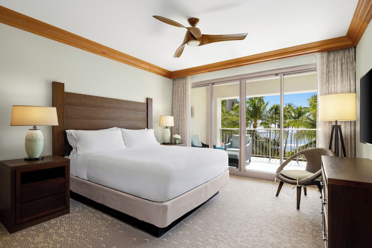 A hotel room with a large bed, two bedside tables with lamps, a ceiling fan, a balcony with a chair, and a TV on a stand.