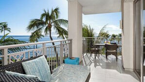 A balcony with wicker furniture, a table, and chairs, overlooking a beach with palm trees and the ocean.