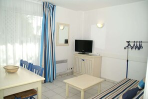 Ein Zimmer mit Fernseher, einem Esstisch mit Stühlen, einer Bank, einem Kleiderständer und einem Fenster mit Vorhängen.