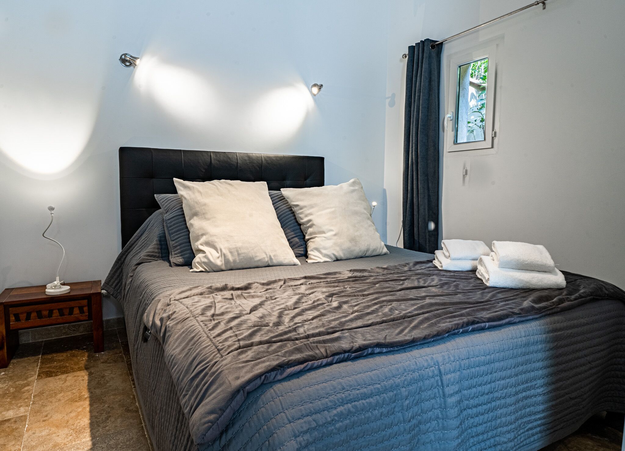 A neatly made bed with a dark headboard, two pillows, and a blue quilt. There is a small bedside table with a lamp, and a window with curtains.
