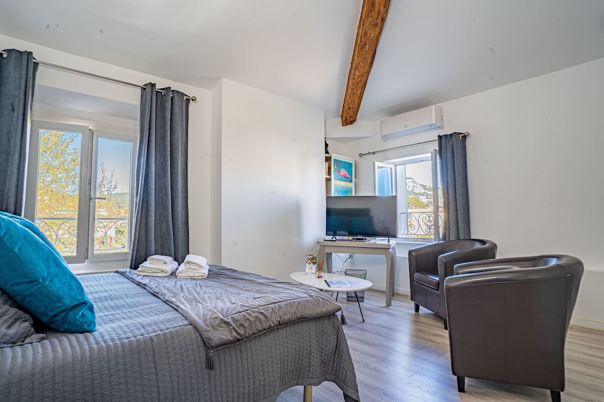 A hotel room with a bed, a desk with a TV, a chair, and a balcony with curtains.