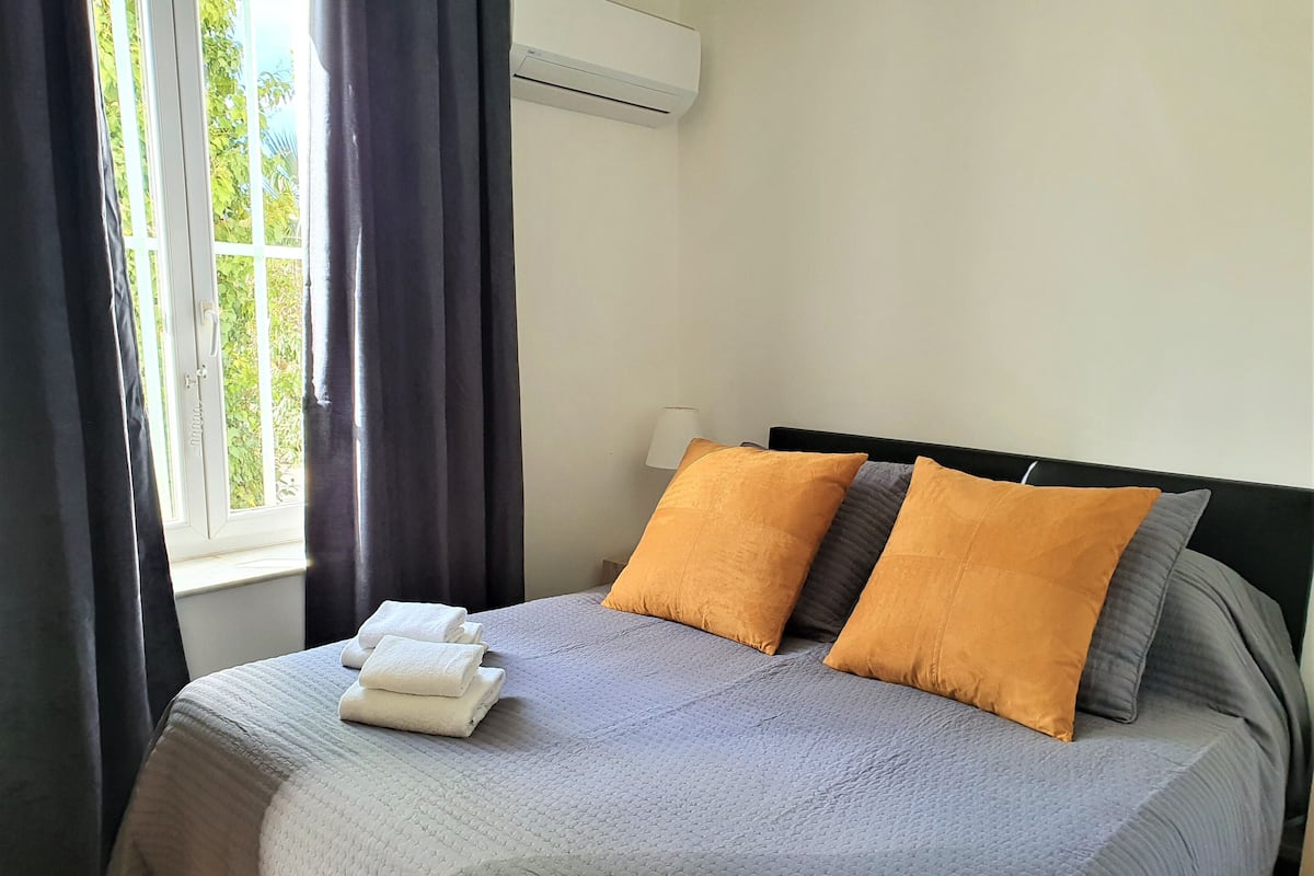 A neatly made bed with a grey quilt, two orange pillows, and a stack of white towels.