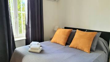 A neatly made bed with a grey quilt, two orange pillows, and a stack of white towels.