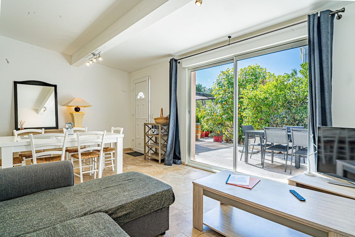 A spacious living area with a sofa, a dining table with chairs, a TV, and a large sliding glass door leading to an outdoor patio with a table and chairs.