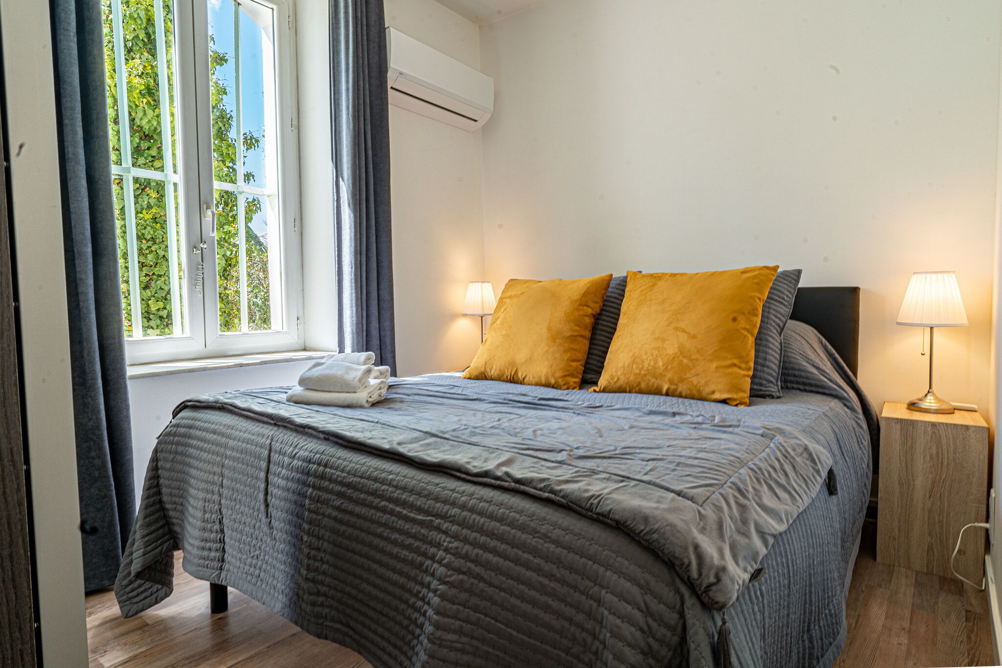 Una cama cuidadosamente hecha con un edredón gris, dos almohadas color mostaza y una toalla blanca al lado. Hay una ventana con cortinas, una unidad de aire acondicionado encima y una lámpara de noche sobre una mesita de madera.