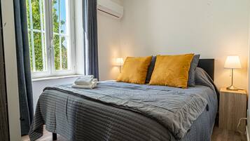 Una cama cuidadosamente hecha con un edredón gris, dos almohadas color mostaza y una toalla blanca al lado. Hay una ventana con cortinas, una unidad de aire acondicionado encima y una lámpara de noche sobre una mesita de madera.
