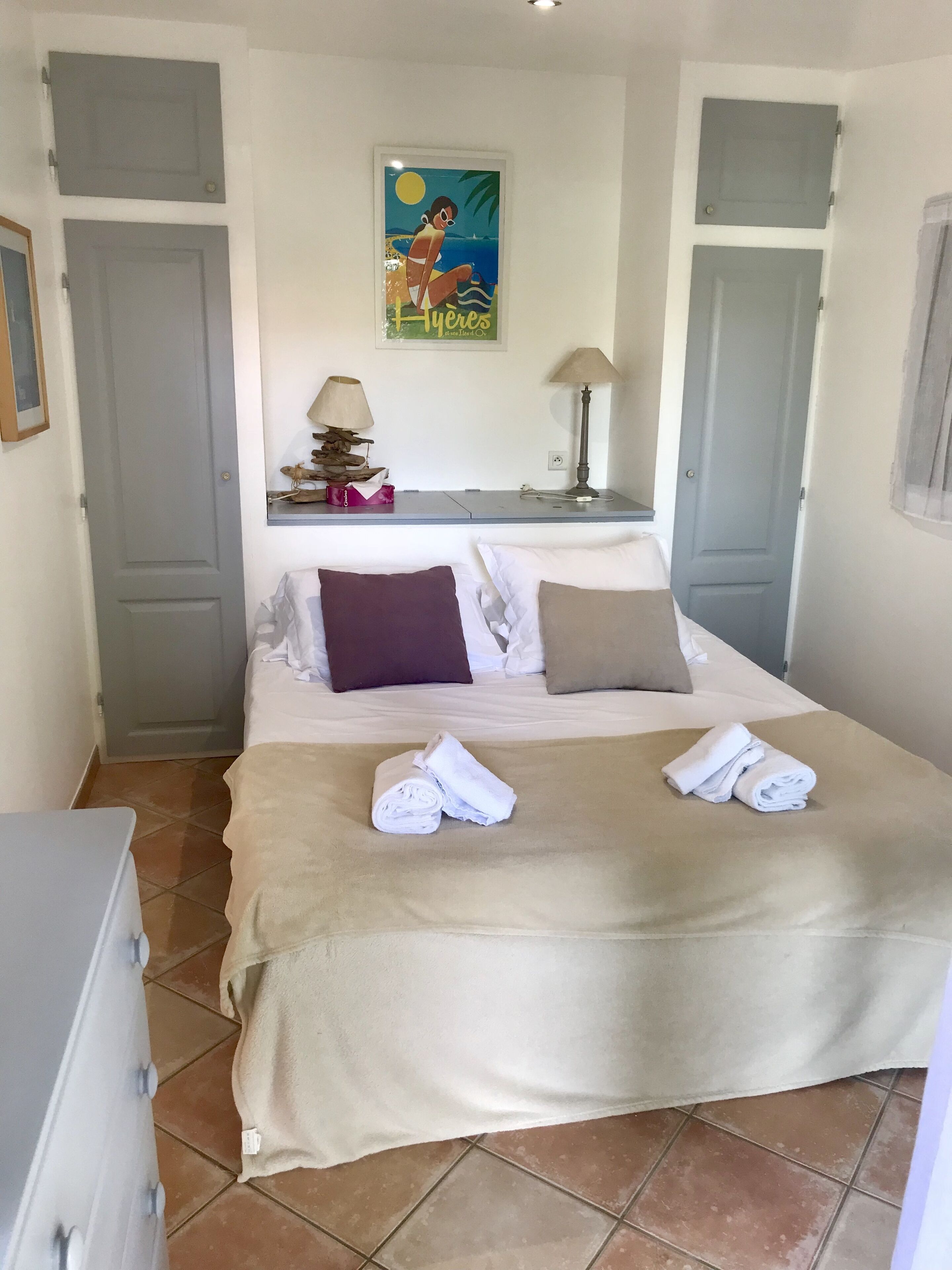 A neatly made bed with a beige blanket, two pillows, and folded towels. There is a small table with a lamp and decorative items. A framed picture hangs on the wall above the bed.
