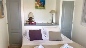 A neatly made bed with a beige blanket, two pillows, and folded towels. There is a small table with a lamp and decorative items. A framed picture hangs on the wall above the bed.