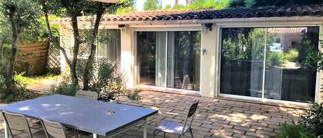 Un patio con mesa y sillas, un espacio de estar cubierto y un ventanal corredizo que da a un área exterior techada.