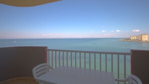 Varanda com uma mesa e cadeiras brancas, com vista para o mar azul e edifícios ao longe.