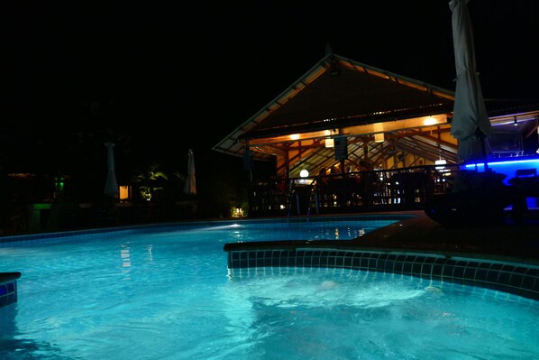 A swimming pool with a tiled edge, a covered seating area with tables and chairs, and a building with a sloped roof in the background.