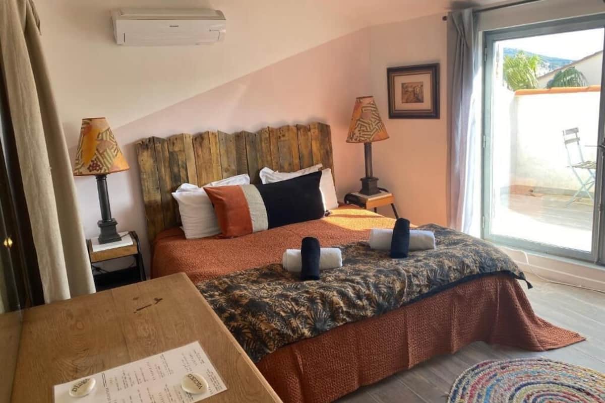 Ein Schlafzimmer mit einem Bett, einem Holztisch, einer Lampe und einem Fenster mit Blick ins Freie.